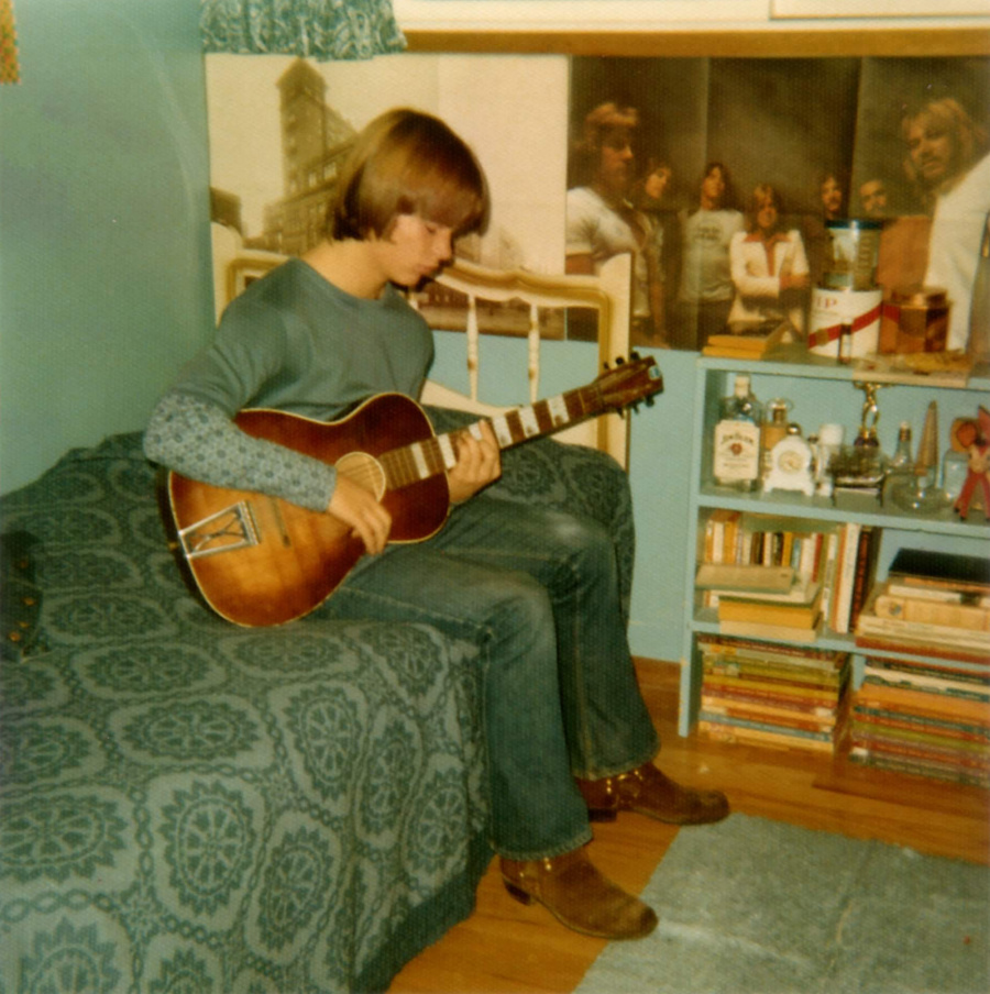 Greg sitting on his bed playing his guitar.